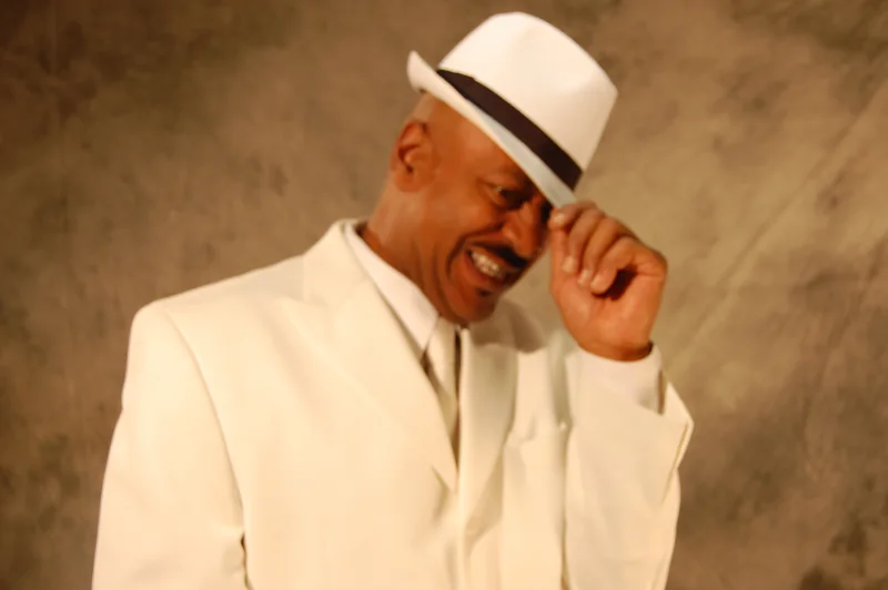 A performer in a white suit and fedora, tipping his hat