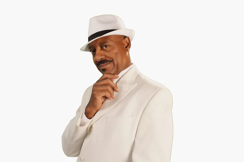 Greg Simmons in a white suit and white fedora, hand to his chin