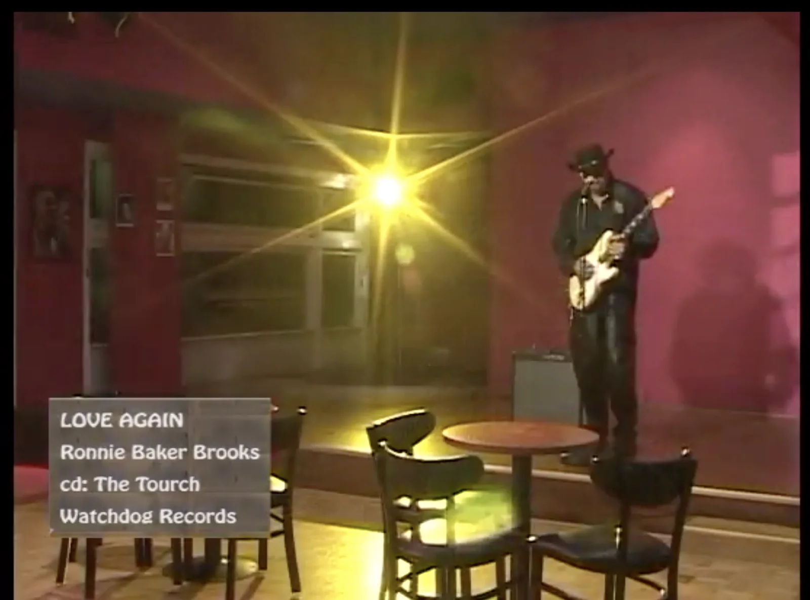 Ronnie Baker Brooks playing guitar on a lit club stage in a frame from a finished music video, with an on-screen caption reading “Love Again”